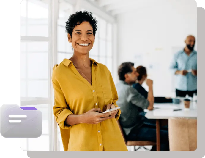 Confident woman in yellow shirt smiling while holding a phone with a team meeting in the background.