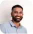 A man with short dark hair and a beard, wearing a light gray collared shirt, smiles while standing against a plain white background, like he’s ready to tackle his Checklist LM tasks for the day.