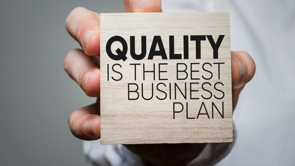 a hand holding a square of wood that says quality is the best business plan