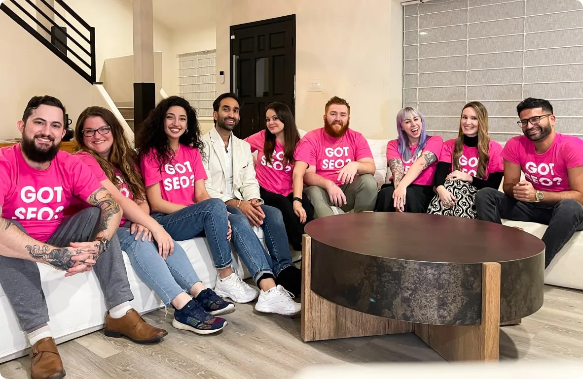 A Search Atlas team of smiling individuals sitting together on a white couch in a modern office lounge, most wearing matching pink 