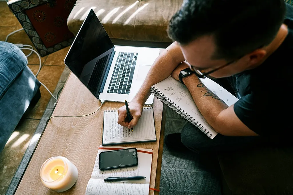 Man writing notes in a notebook while working on a laptop at home with a candle and phone on the table