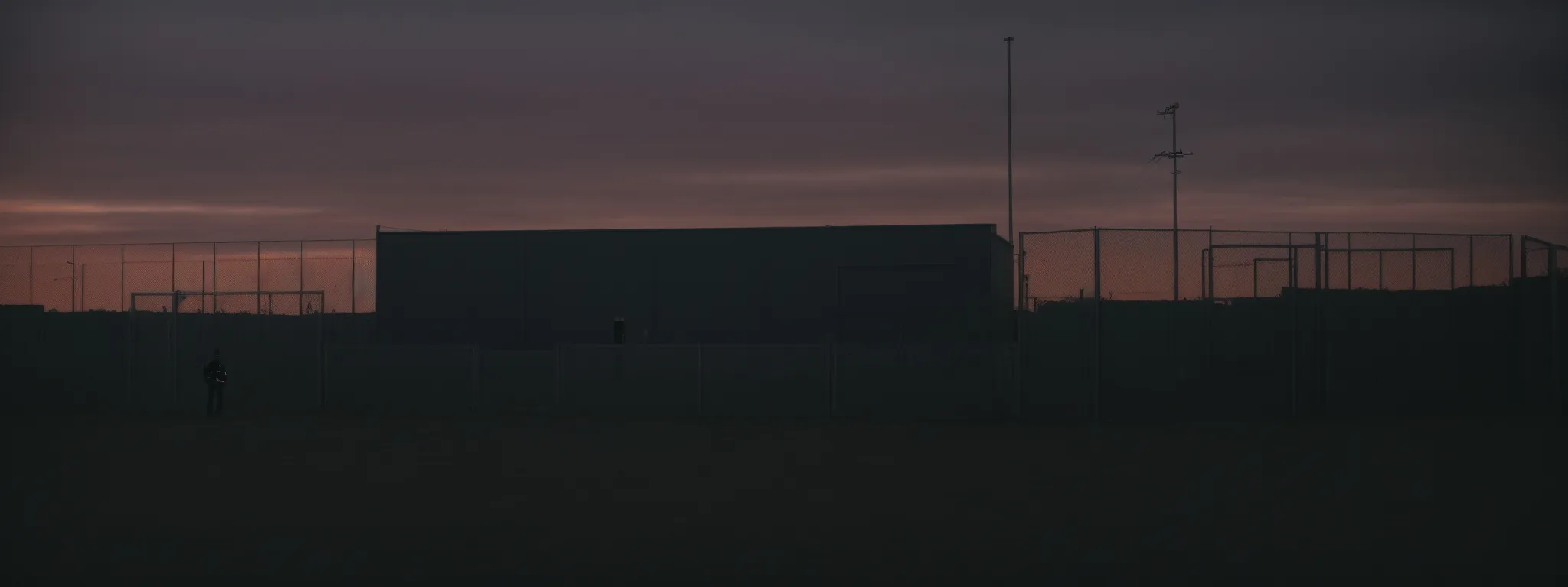 a security guard patrols the perimeter of a fenced and gated server farm at dusk.