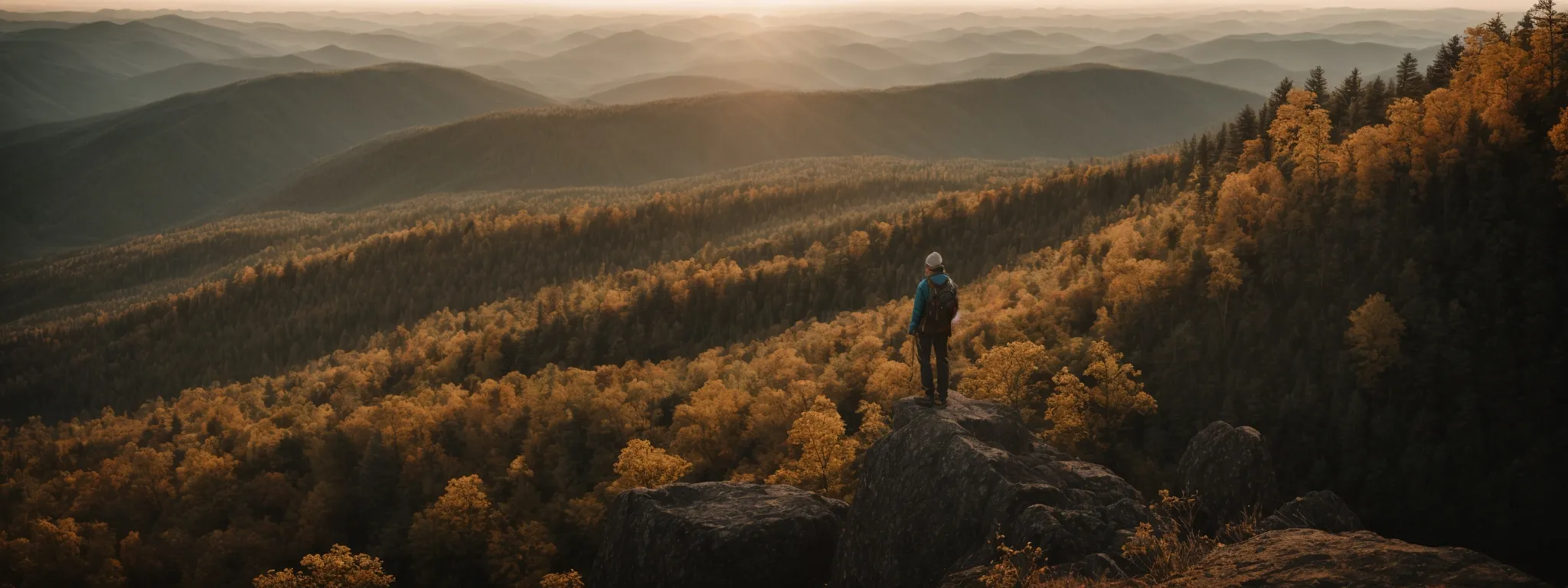 a person stands atop a mountain peak, looking out over a vast forested landscape at sunrise.