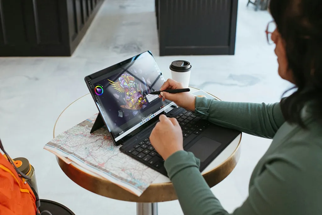 Woman sketching digital artwork on a touchscreen tablet with stylus in a cafe setting, map and coffee on table