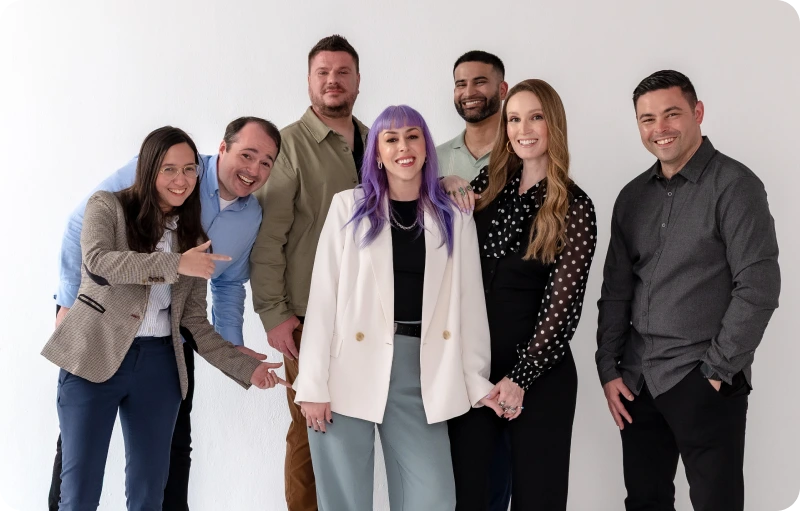 Seven adults stand and smile together in front of a white wall, with one person in the center wearing a white blazer and purple hair, representing an innovative Enterprise SEO Software team.