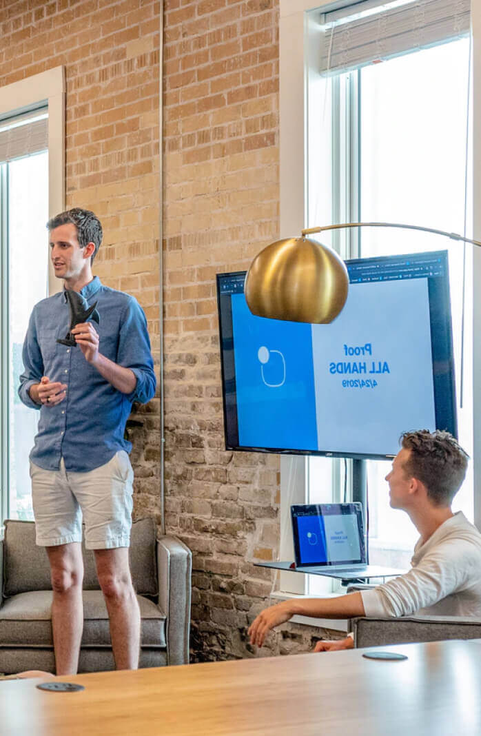 A person stands and presents with a phone in hand next to a screen displaying “Proof All Hands” while another person sits nearby in a modern office setting.