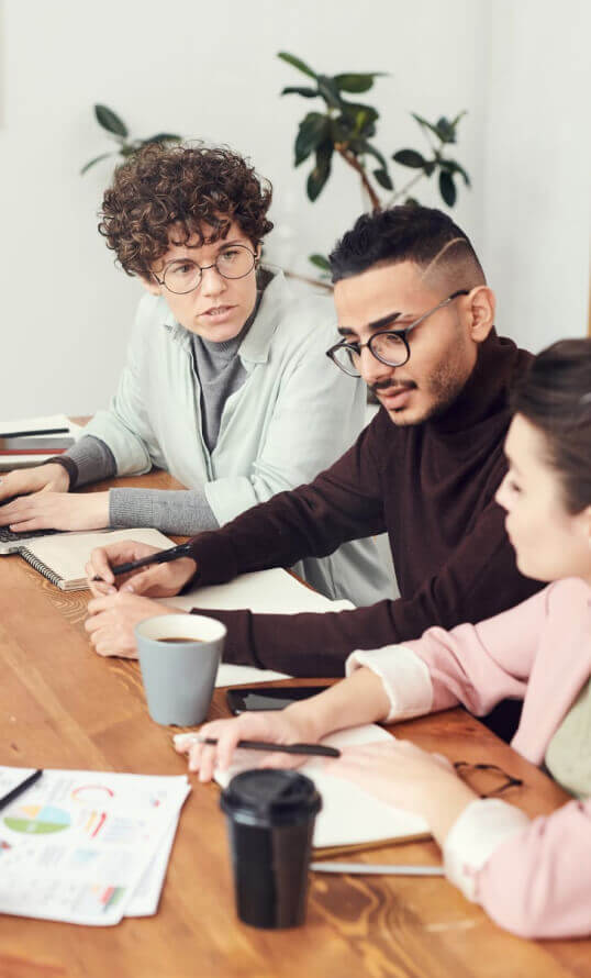 Three people sit at a wooden table with notebooks, papers, and coffee cups, engaged in discussion.