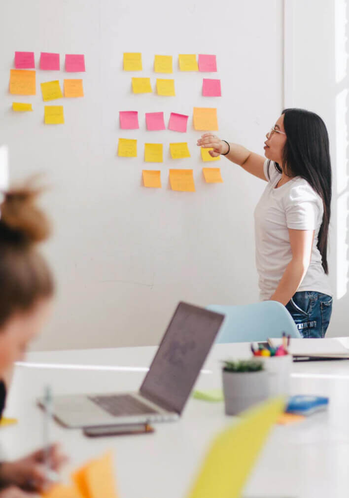 A woman stands at a whiteboard covered with colorful sticky notes, organizing them for an SEO Reputation Management strategy, while another person works at a desk with a laptop in the foreground.