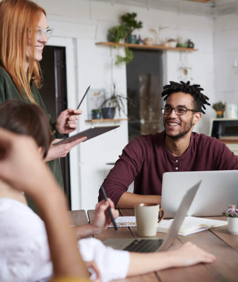 Four people sit at a table with laptops and notebooks, engaged in a discussion about SEO Reputation Management. One person stands holding a tablet, while another smiles and listens. Indoor office setting with plants.